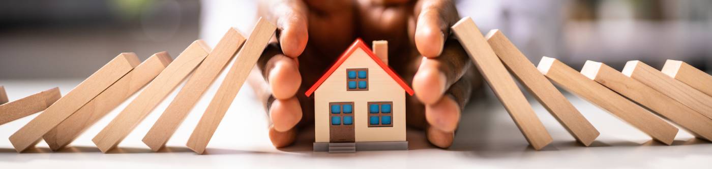 small wooden house being protected by hands as dominos fall toward it